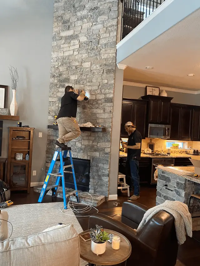 G4 Electric technician on a step ladder wiring above a stacked-stone fireplace in a living room
